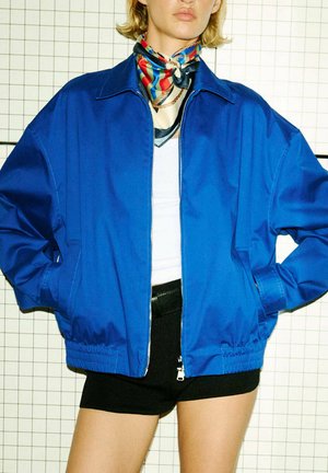 Persona con el pelo corto y rubio lleva una chaqueta azul con cremallera, top blanco, pantalones cortos negros y un pañuelo de cuello colorido con estampado frente a un fondo cuadriculado.