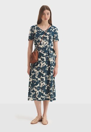 Bloemenjurk in marineblauw en crème, met een V-hals, korte mouwen en een tailleband. Gecombineerd met een bruine crossbodytas en beige schoenen.