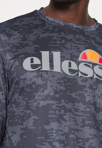 Ciemnoszary sportowy t-shirt z pikselowym wzorem, okrągłym dekoltem oraz logo "ellesse" w jasnoszarym kolorze z kolorowym półsłonecznym akcentem.