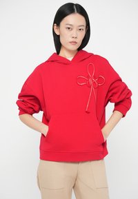 Νεαρός άνθρωπος με ίσια μαύρα μαλλιά, φορώντας μια κόκκινη φούτερ με διακοσμητική λεπτομέρεια στα κορδόνια και μπεζ παντελόνι, στέκεται με τα χέρια στις τσέπες.