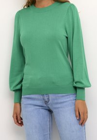 Femme portant un pull vert à manches longues bouffantes et un jean bleu clair, vue des épaules jusqu'aux hanches sur un fond uni.