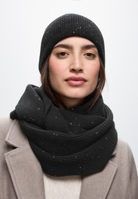 Sort strikket beanie og oversized tørklæde med glitrende detaljer, tekstureret mønster og lagdelt design, båret over en lys sweater og frakke.