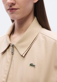 Μπεζ ζακέτα με φερμουάρ, με ριμπ ύφασμα στο γιακά και μικρό πράσινο λογότυπο κροκόδειλου κεντημένο στο αριστερό στήθος. Με λεία υφή υφάσματος.