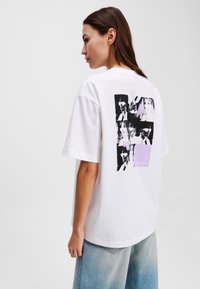 White cotton t-shirt featuring a graphic print on the back with black and grey images, accented by a small lavender rectangle. Boxy fit.