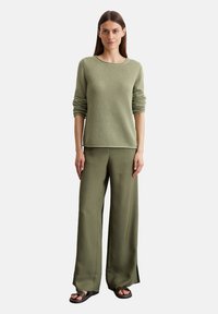 Maglione verde chiaro, texturizzato, con maniche arrotolate abbinato a pantaloni larghi verde oliva e sandali neri, in piedi su uno sfondo bianco.