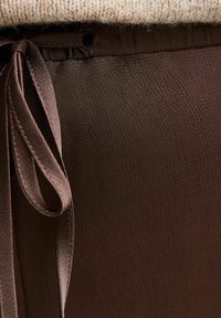 Brown satin fabric with a smooth texture, featuring a tied ribbon detail at the waist and visible stitching along the edges.