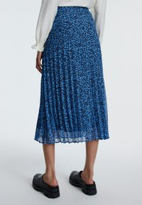 Faltender Midirock in kräftigem Blau mit einem schwarzen Blumenmuster. Hergestellt aus leichtem Stoff. Kombiniert mit klobigen schwarzen Schlupfschuhen.
