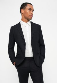 Μαύρο σακάκι με ταίριασμα, με σχέδιο μονής αναγκαίας, δύο κουμπιά, πέλματα λεπίδας και τσέπη στήθους, συνδυασμένο με λευκό πουκάμισο.