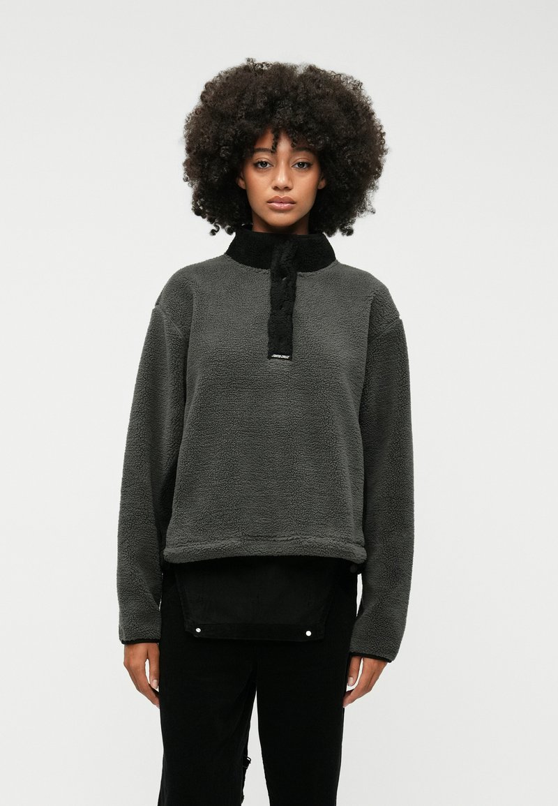 Γκρίζος φλις πουλόβερ με ψηλό γιακά και μαύρες λεπτομέρειες. Διαθέτει μπροστινό φερμουάρ, άνετη εφαρμογή και υφή υφάσματος. Μαύρο παντελόνι από κάτω.