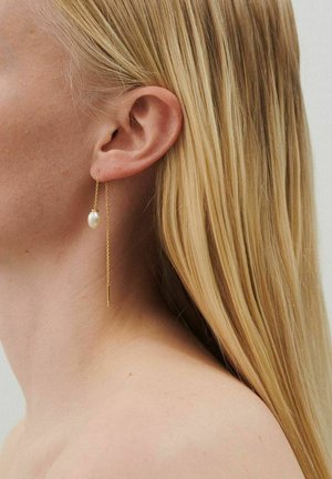 Primer plano de la oreja de una mujer con cabello rubio largo, que lleva un delicado pendiente de cadena de oro con una pequeña perla blanca.