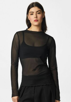 Jeune femme aux cheveux foncés portant un haut noir transparent à manches longues et un pantalon noir, debout devant un fond clair uni.
