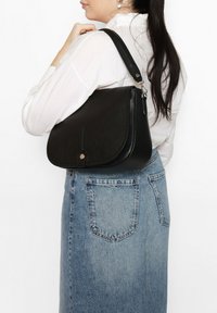 Zwarte leren schoudertas met een ronde klep, een gestructureerde afwerking en een gouden accent. Draag aan de zijkant met een spijkerrok.