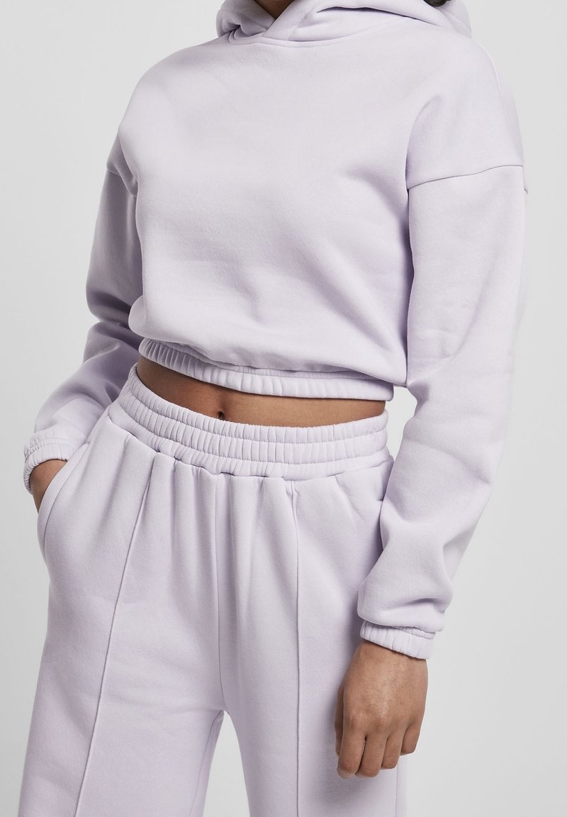 Femme portant un sweat à capuche court violet clair et un pantalon de survêtement taille haute assorti, les mains dans les poches, sur un fond uni.