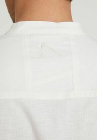 Camicia bianca con un design pulito, tessuto leggero e un sottile dettaglio ricamato vicino al colletto sul retro.