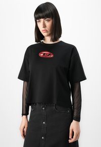 Ung kvinne med rett, mørkt hår iført lagdelt svart topp med rødt logo og knappet svart denimskjørt, stående mot hvit bakgrunn.