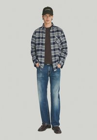 Camicia di flanella a quadri in blu, bianco e grigio sopra una t-shirt marrone; jeans blu; berretto nero con testo bianco; scarpe marroni.