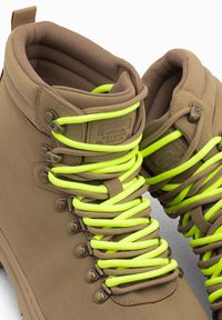 Botas de senderismo marrones con cordones amarillo neón, acabado mate, ojales de metal y cuellos acolchados para comodidad y soporte.