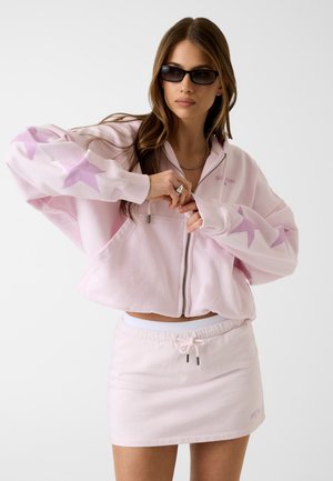 Frau trägt hellrosa Kapuzenjacke mit Sternenmuster und passenden Minirock, zieht den Reißverschluss der Kapuzenjacke hoch, mit langen braunen Haaren und dunkler Sonnenbrille.