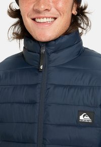 Usmievajúca sa osoba s jemnými pehami, oblečená v tmavomodrom prešívanom kabáte so zapnutým zipsom, na hrudi s logom Quiksilver.