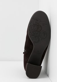 Semelle de bottine en daim marron foncé avec logo embossé, motifs de pièces de monnaie et talon bloc reposant contre une surface d'angle blanche.
