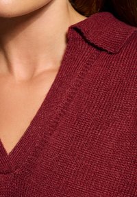 Burgundowa sweter robiony na drutach z dekoltem w kształcie litery V i kołnierzykiem, z teksturowanym materiałem i widocznymi ściegami wzdłuż linii dekoltu.