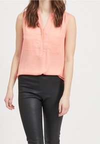 Blouse sans manches de couleur pêche avec un col en V, dotée de deux poches poitrine. Associée à des leggings en cuir noirs et ajustés. Texture lisse.