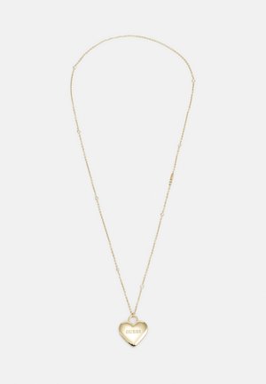 Collier délicat en chaîne d'or avec de petites perles et un pendentif en forme de cœur gravé du mot « GUESS » sur un fond blanc.