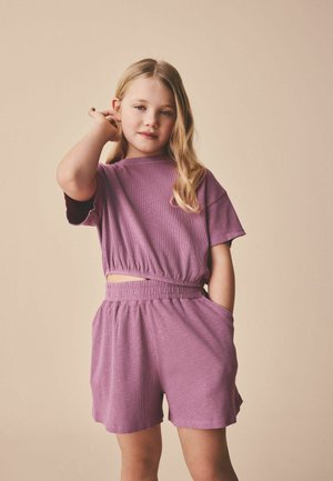 Ung pige med langt blondt hår iført en matchende mauve ribbet kortærmet top og shorts, poserer med den ene hånd i lommen og den anden rører ved håret.
