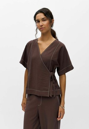 Femme aux cheveux bruns attachés, portant un haut cache-cœur texturé marron à manches courtes et un pantalon assorti sur un fond blanc uni.