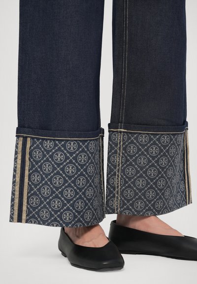 Džinsa bikses ar rakstainu aproci, kurai ir ģeometrisks raksts tumši zils, zelta svītras un kontrastējošas šuves. Melni dzīvokļi papildina izskatu.