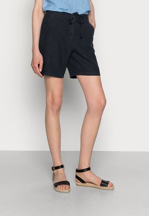 Vrouw draagt zwarte shorts met trekkoord, lichtblauw shirt en zwarte sandalen met enkelbandje, staand op een grijze vloer tegen een witte muur.