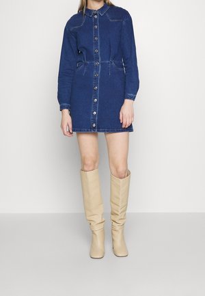 Mujer con un mini vestido vaquero azul abotonado, de manga larga, y botas beige arrugadas hasta la rodilla, sobre un fondo liso.