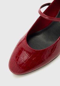 Chaussure Mary Jane en cuir verni rouge avec texture crocodile et semelle intérieure noire sur un fond clair, montrant le devant et le détail de la bride.