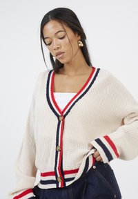 Cardigan lavorato a maglia color crema, con strisce rosse e blu navy, scollo a V profondo e bottoni in legno. Vestibilità ampia con polsini e orlo a coste.