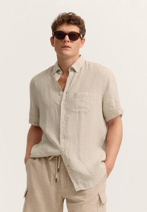 Mann trägt beiges kurzärmeliges Hemd mit Knöpfen, passende beige Cargoshorts mit Kordelzug und dunkle Sonnenbrille, steht mit Händen in den Taschen.