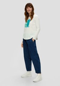 T-shirt blanc à manches longues avec un imprimé abstrait bleu, associé à un jean bleu foncé ample et des baskets blanches à plateforme.