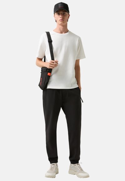 Černé tepláky, bílé tričko s krátkým rukávem, černá čepice s logem a černá crossbody taška. Béžové tenisky s texturovanou podrážkou.