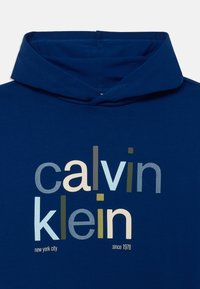 Plava dukserica s grafičkim printom na kojem se nalazi natpis "calvin klein" u različitim fontovima i bojama, a ispod piše "new york city since 1978".