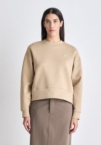 Sudadera de algodón beige con cuello redondo, puños y dobladillo de canalé, que presenta un pequeño logo en relieve en el pecho. Textura suave.