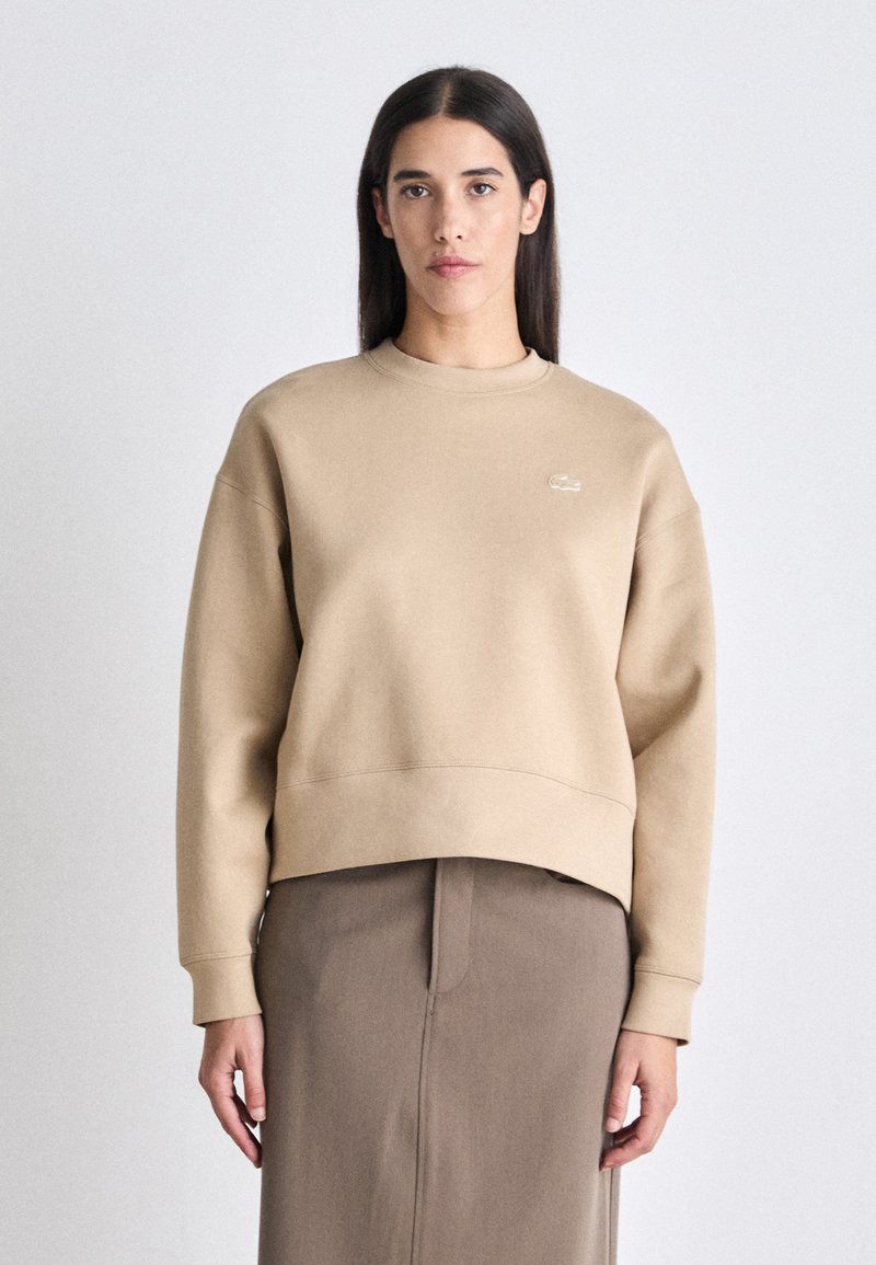 Sudadera de algodón beige con cuello redondo, puños y dobladillo de canalé, que presenta un pequeño logo en relieve en el pecho. Textura suave.