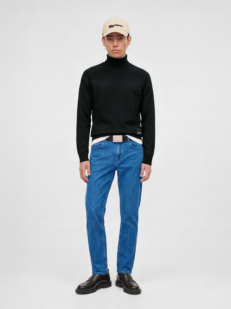 Jeune homme portant une casquette beige, un pull à col roulé noir, un jean bleu, des chaussures noires, debout face à l'avant sur un fond uni.