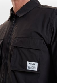 Černá lehká bunda se zipem vpředu, dvěma náprsními kapsami, skládacím límcem a bílou nálepkou na levé kapse s podrobnostmi o značce.