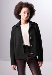 Giacca nera in misto lana con colletto, chiusura con doppio petto e dettagli sulle spalle, abbinata a un maglione chiaro e pantaloni bordeaux.
