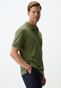 Polo shirt verde oliva con colletto standard, maniche corte e orlo dritto, abbinato a pantaloni blu scuro. Tessuto morbido.