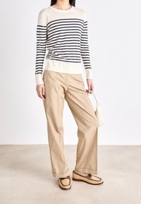 Sweter w paski w kolorze ecru i granatowym, beżowe spodnie i plecione mokasyny z brązowymi akcentami, trzymając małą białą torebkę.