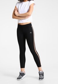 Černé sportovní legíny s růžovými bočními pruhy, spárované s bílým zkráceným topem s malými otvory pro prodyšnost. Šedé tenisky.