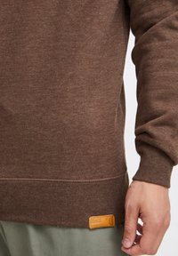 Sudadera marrón con puños y dobladillo de canalé, que presenta un tejido texturizado y una etiqueta de cuero rectangular en el lado inferior.