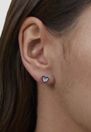 Ohrringe in Herzform mit silberner Umrandung und perlmuttartiger Innenfläche, vor neutralem Hintergrund präsentiert. Ohr teilweise sichtbar.