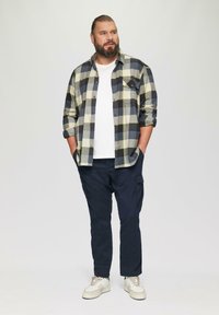 Herenoutfit bestaande uit een zwart en gele geruite flanellen overhemd over een witte ronde hals t-shirt, gecombineerd met marineblauwe cargobroeken en witte sneakers.