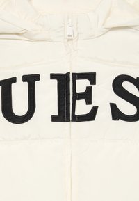 Balta pūsta striukė su didelėmis juodomis siuvinėtosiomis raidėmis, rašančiomis "QUEST" ant priekinės dalies, su užtrauktuku ir gobtuvu.